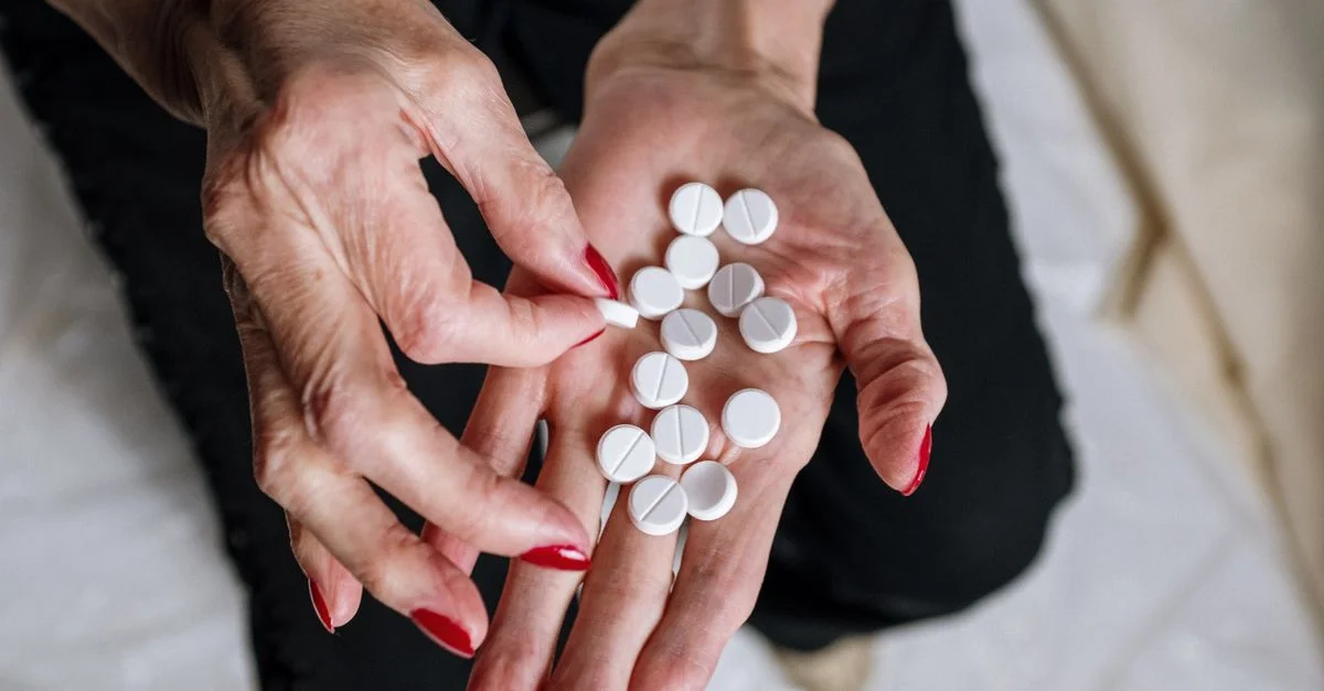 Les mains d'une femme âgée avec des ongles rouges tenant des comprimés ronds blancs, représentant le concept de médication.
