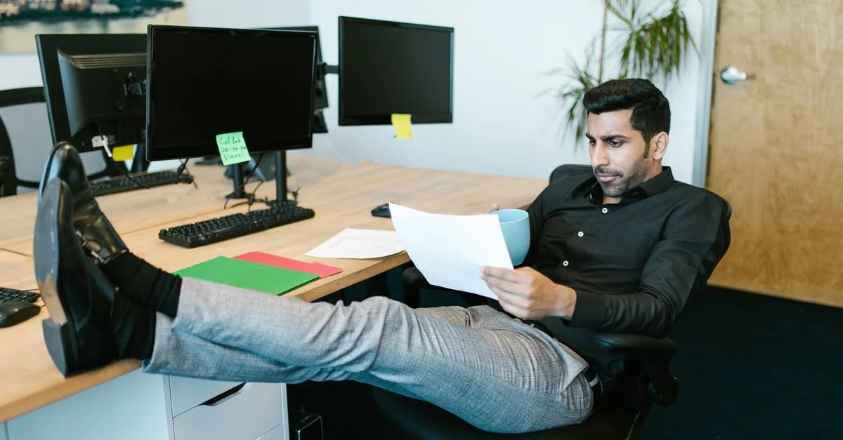 Employé de bureau se détendant et consultant des documents tout en tenant une tasse de café, les pieds sur le bureau.