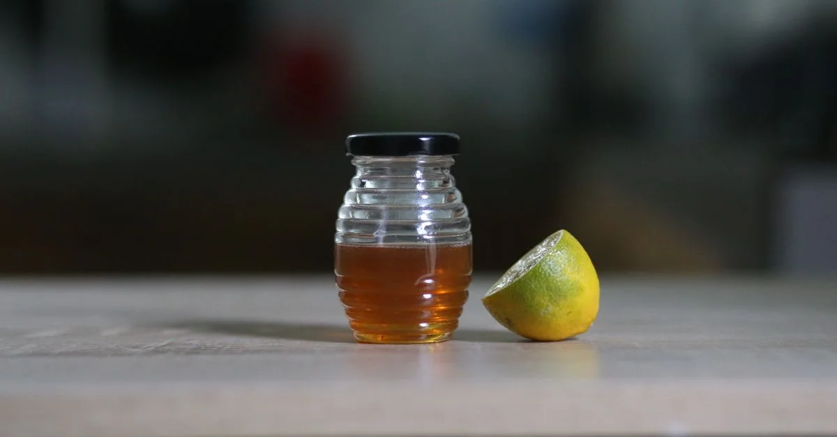 Un pot en verre rempli de miel à côté d'un citron tranché sur une table en bois à l'intérieur.