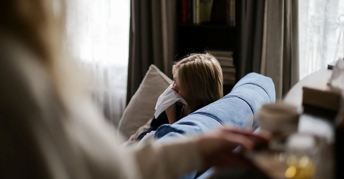 Un jeune enfant est assis sur un canapé à l'intérieur, utilisant un mouchoir, suggérant une maladie ou une allergie.