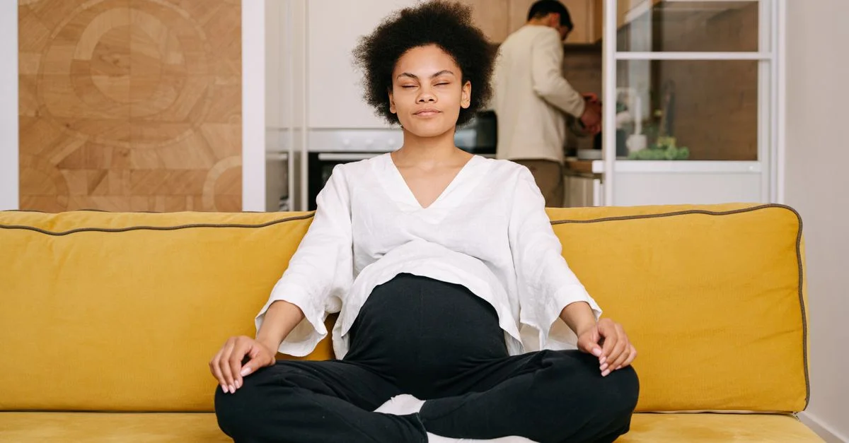 Femme enceinte méditant sur un canapé jaune dans un cadre de maison douillette, mettant l'accent sur la relaxation et le soin de soi.