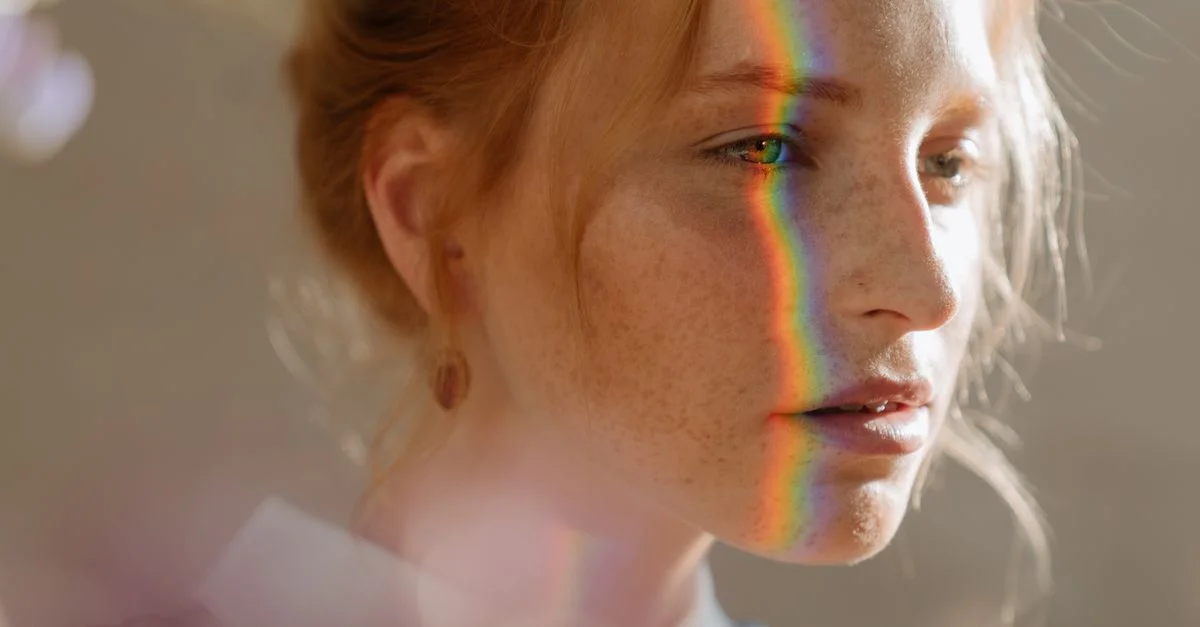 Femme rousse aux taches de rousseur avec des lumières arc-en-ciel sur le visage, créant un effet magique et rêveur.