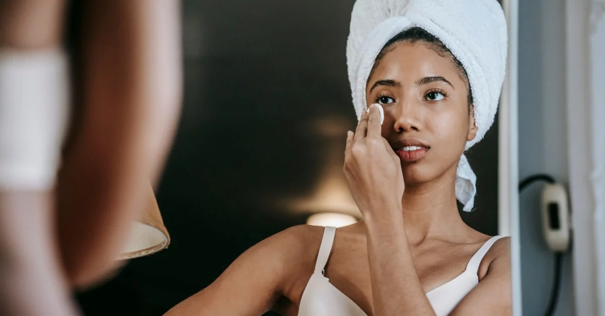 Femme adulte appliquant des soins de la peau tout en se regardant dans un miroir, portant une serviette sur la tête.