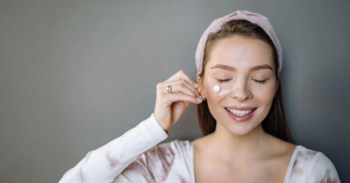 Femme souriante appliquant une crème skincare les yeux fermés sur fond gris.