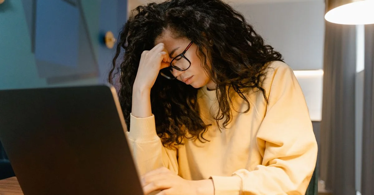 Une femme fatiguée avec des cheveux bouclés et des lunettes ressentant du stress en travaillant sur un ordinateur portable.