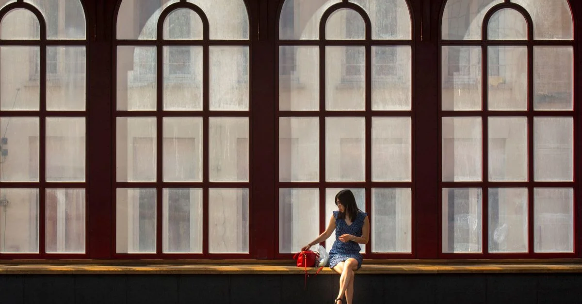 Une femme est assise seule dans la gare d'Anvers, silhouettée par de grandes fenêtres en arc.