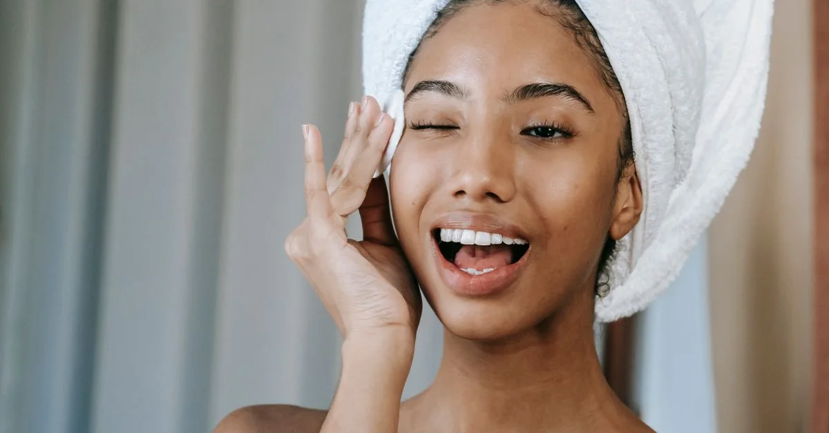 Jeune femme avec une serviette sur la tête, souriant et appliquant un produit de soins de la peau, promouvant une routine de beauté matinale.