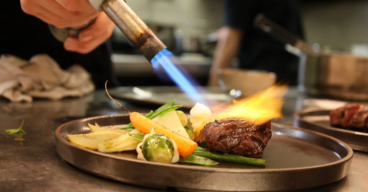 Saisir un plat gourmet avec des légumes frais à l'aide d'un chalumeau culinaire dans une cuisine professionnelle.