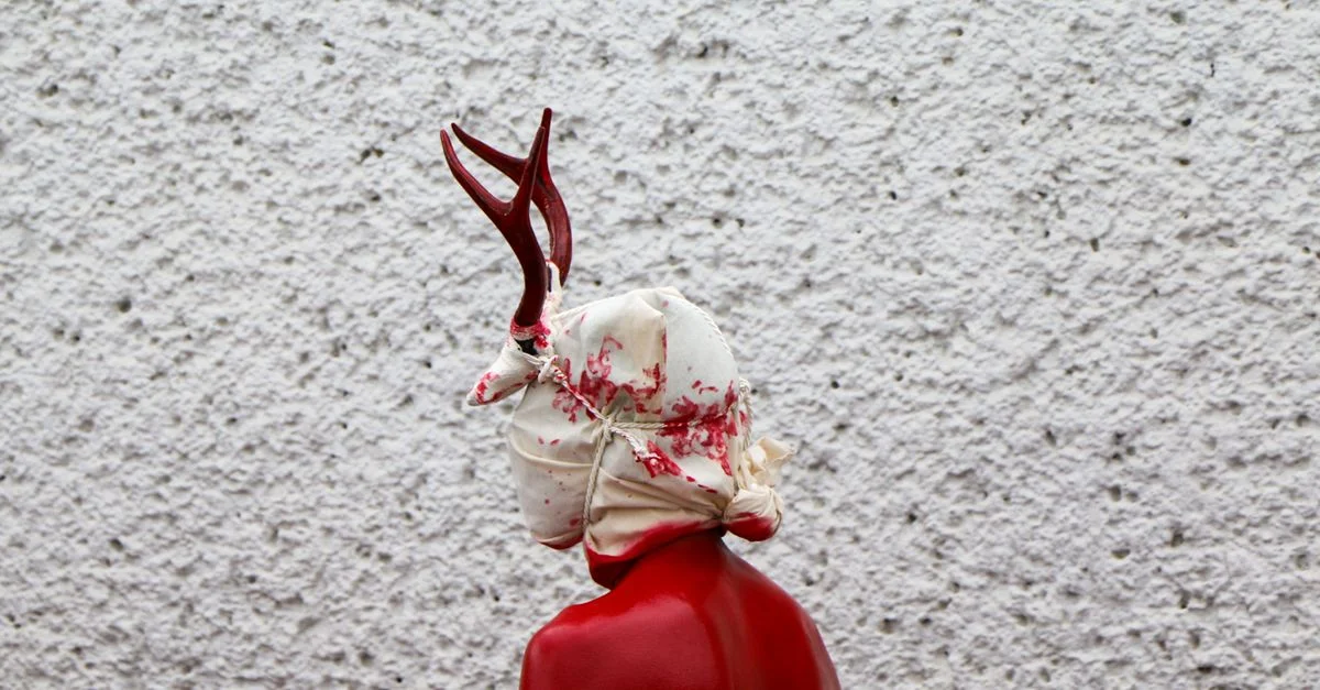 Vue de profil d'une figure peinte en rouge avec des bois de cerf et un masque ensanglanté, devant un mur texturé.