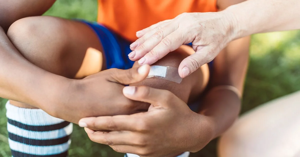Une vue en gros plan d'un adulte appliquant un pansement sur le genou blessé d'un enfant en extérieur pendant la journée.