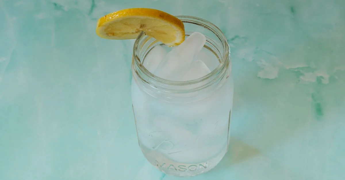 Gros plan sur une eau glacée rafraîchissante dans un bocal en verre avec une tranche de citron sur un fond turquoise, idéal pour des thèmes d'été.