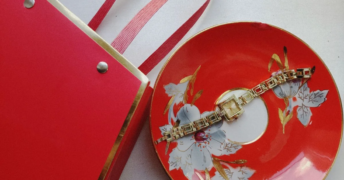 Élégante montre-bracelet en or posée sur une assiette décorative rouge à motifs floraux, à côté d'une boîte cadeau.