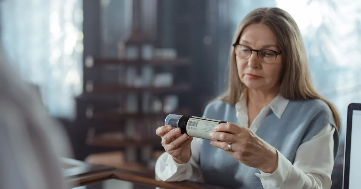 Une femme senior avec des lunettes examine une bouteille de médicament au comptoir d'une pharmacie à l'intérieur.