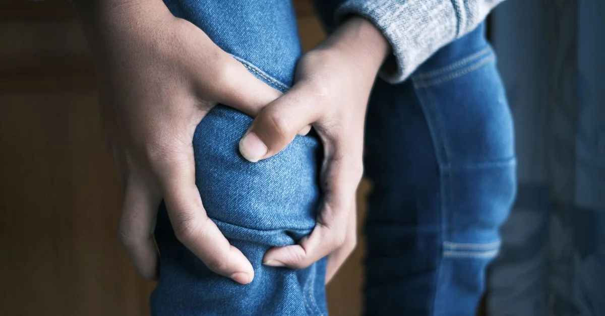 Image en gros plan de mains tenant un genou dans un jean, indiquant de la douleur ou une blessure.