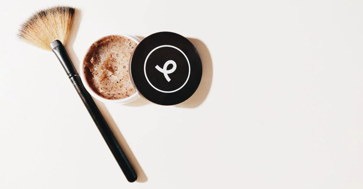 Vue en plongée d'un pinceau de maquillage et d'un récipient à gommage sur une surface blanche, parfait pour les thèmes de beauté et de soins de la peau.