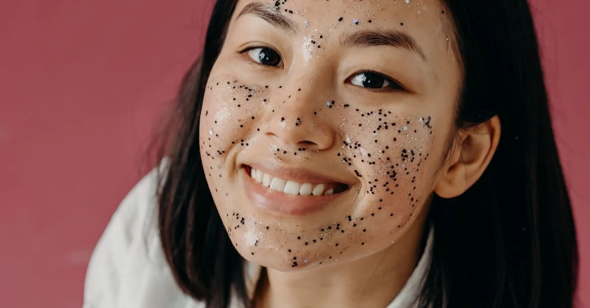 Femme asiatique souriante avec un masque facial pailleté sur fond rose, promouvant les soins de la peau et la beauté.