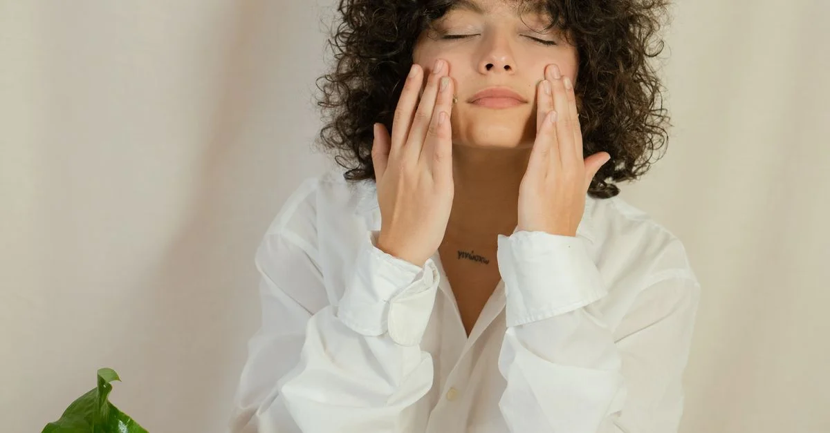 Femme caucasienne aux cheveux bouclés appliquant délicatement de la crème pour le visage pour une routine de soin relaxante à l'intérieur.