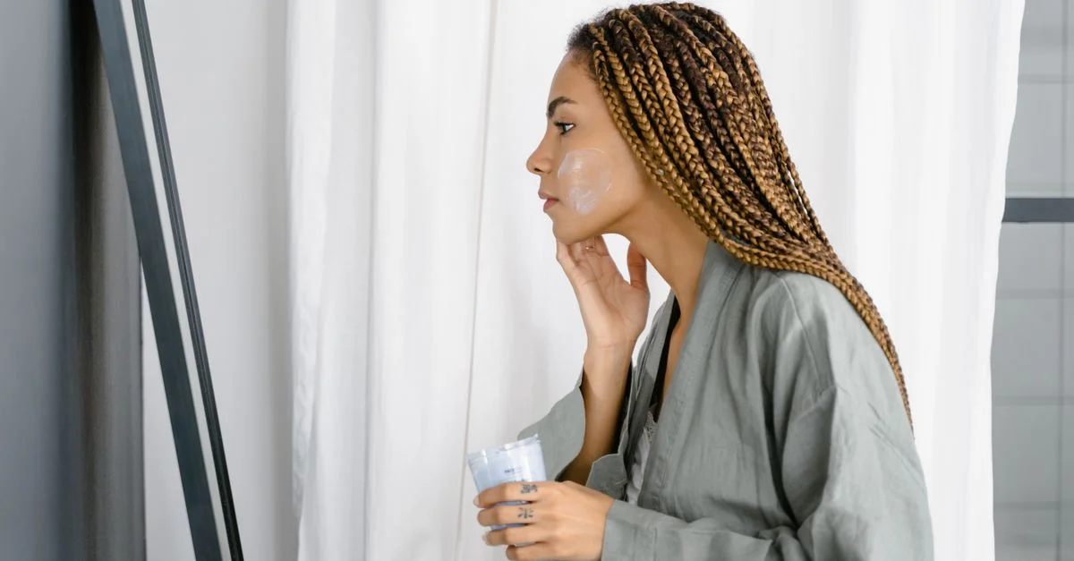 Une femme en robe verte appliquant une crème de soins sur son visage devant un miroir à l'intérieur.