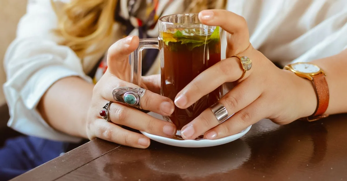 Gros plan sur une femme tenant du thé herbal dans un verre, ornée de bagues et d'une montre.