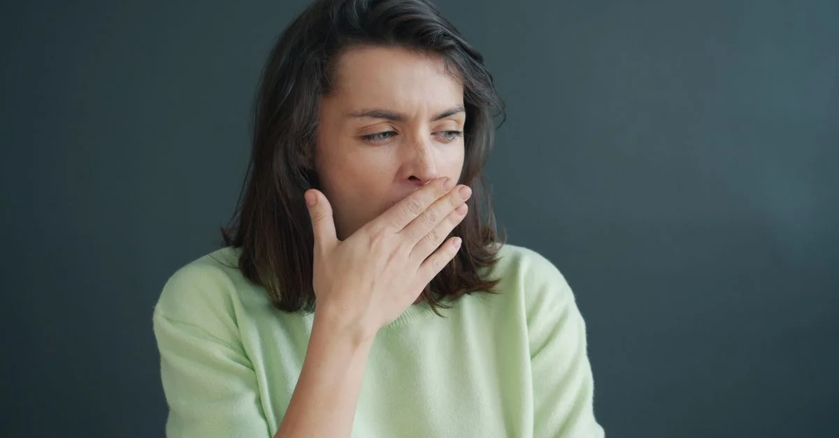 Jeune femme bâillant, la main sur la bouche, vêtue d'un pull décontracté, sur fond gris, en intérieur.