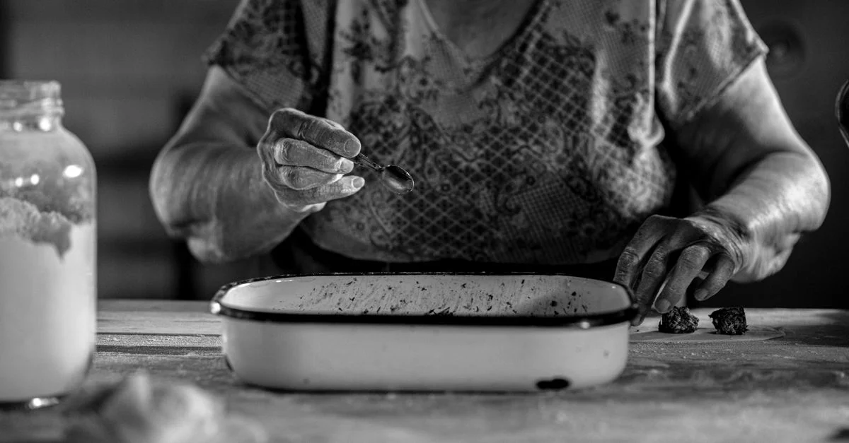Photo en noir et blanc d'une femme âgée préparant des biscuits, mettant l'accent sur la cuisine traditionnelle.