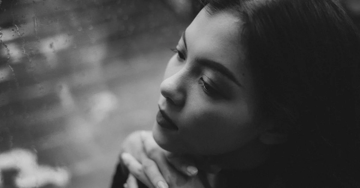 Portrait en noir et blanc d'une femme adulte regardant par une fenêtre sous la pluie, plongée dans ses pensées.