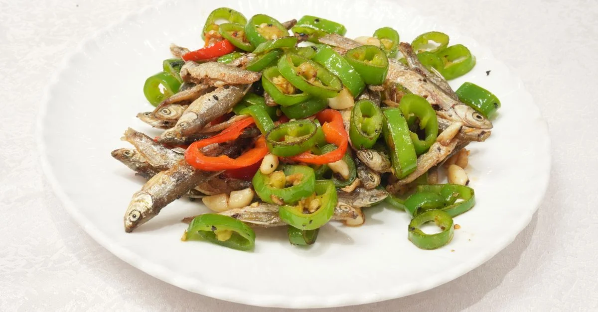 Un plat d'anchois frits accompagnés de poivrons verts et rouges émincés, servi sur une assiette blanche.