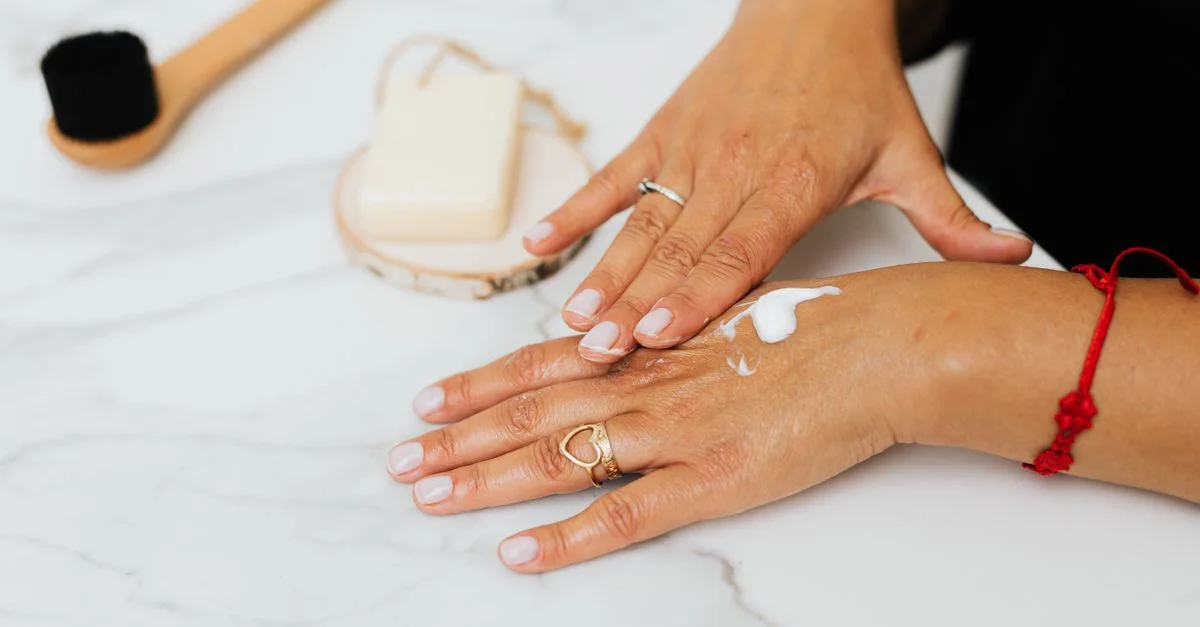 Gros plan sur une femme appliquant une crème hydratante sur ses mains avec des produits de soin à proximité.