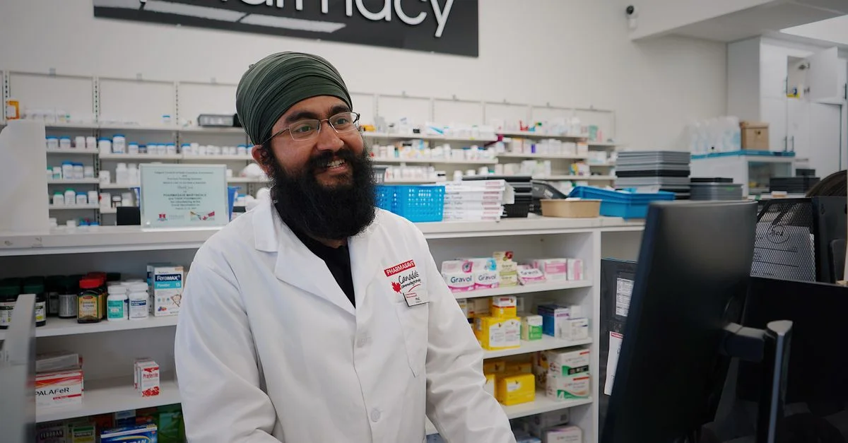 Pharmacien masculin avec un turban se tenant et souriant dans une pharmacie, travaillant au comptoir.