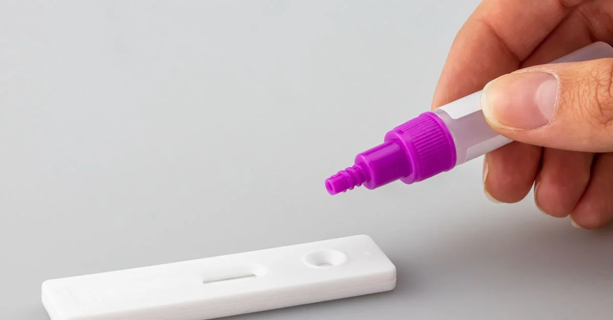 Gros plan sur une main appliquant du liquide sur un kit de test rapide, sur une surface blanche.