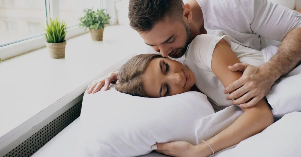 Un couple amoureux, enlacé dans un lit près d'une grande fenêtre, savoure son intimité.