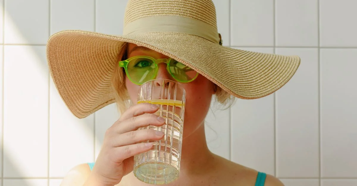 Femme adulte portant des lunettes de soleil vertes, buvant de l'eau avec une tranche de citron, portant un chapeau.