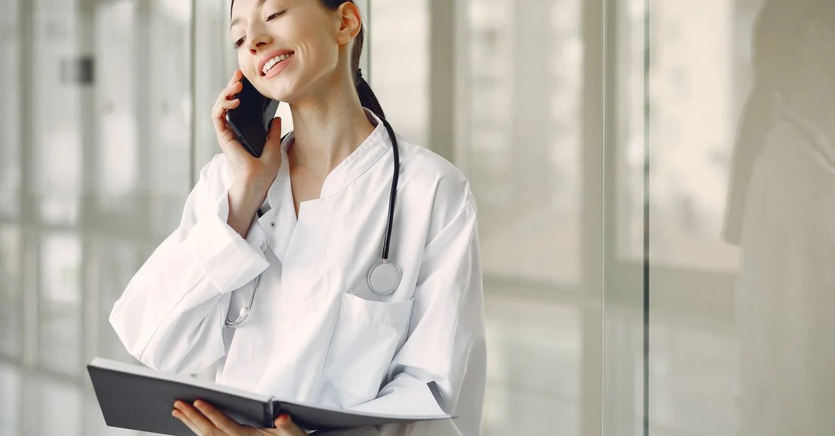 Médecin féminin amical en blouse blanche parlant au téléphone portable, tenant un carnet dans une clinique.