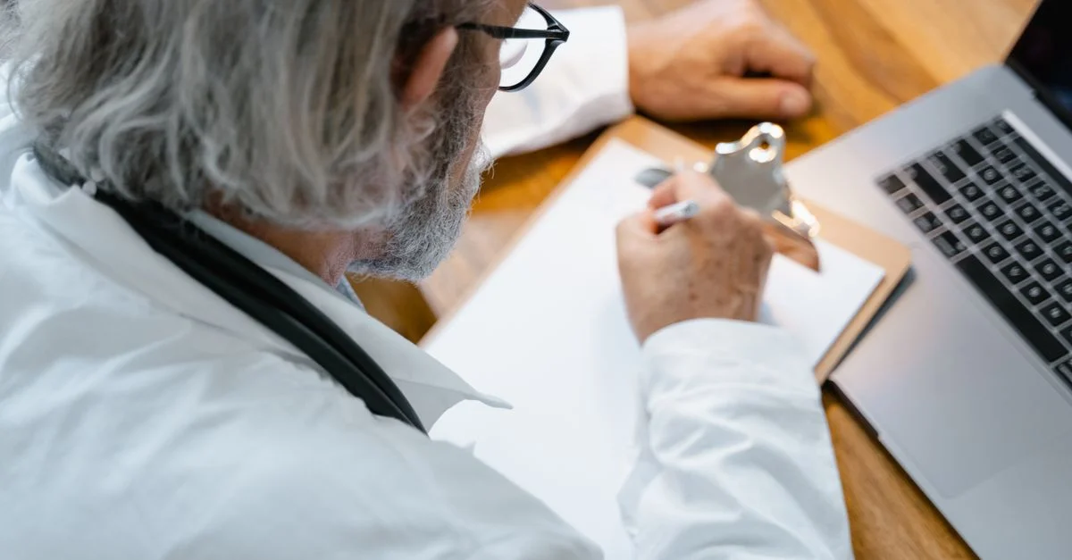 Un médecin âgé en blouse blanche prenant des notes sur un cahier à un bureau avec un ordinateur portable.