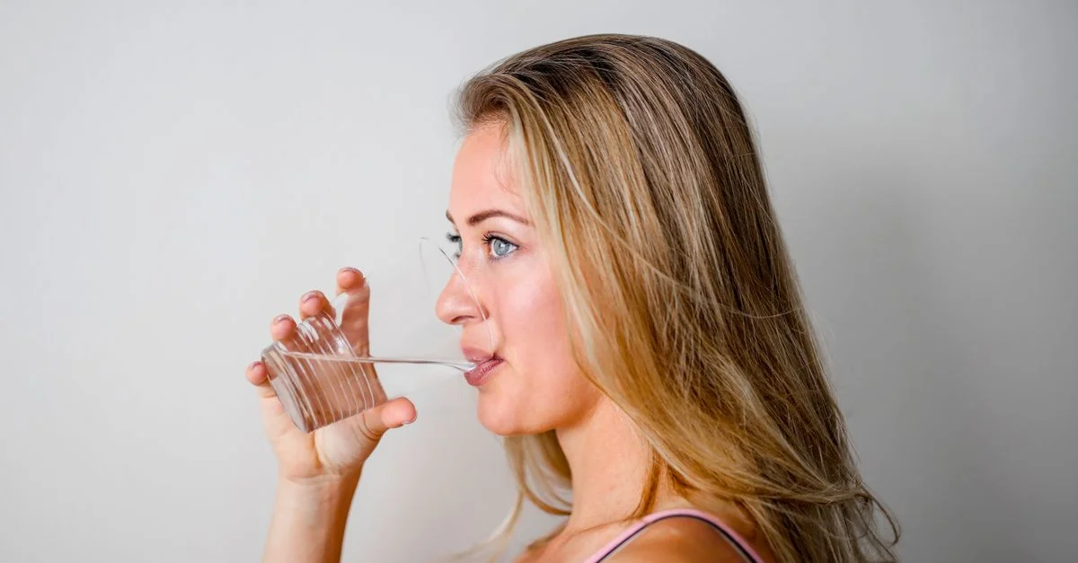 Vue de côté d'une femme aux cheveux blonds buvant de l'eau dans un verre transparent.