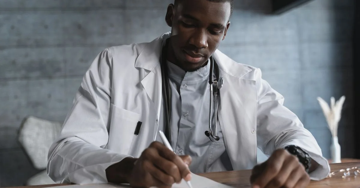 Médecin noir en blouse de laboratoire prenant des notes à un bureau en bois dans un cabinet médical.