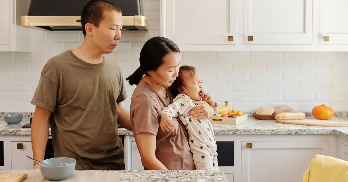 Une famille asiatique réconforte un bébé qui pleure dans une cuisine moderne, illustrant la parentalité et le soin.