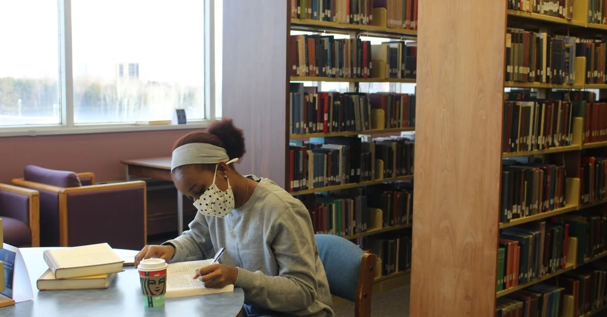 Jeune femme dans une bibliothèque portant un masque, concentrée sur ses études.