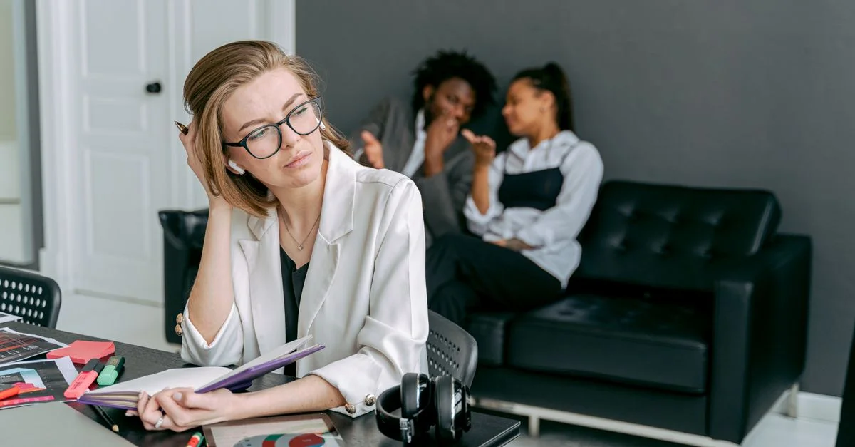 Une femme semble stressée à son bureau, avec des collègues qui murmurent en arrière-plan.