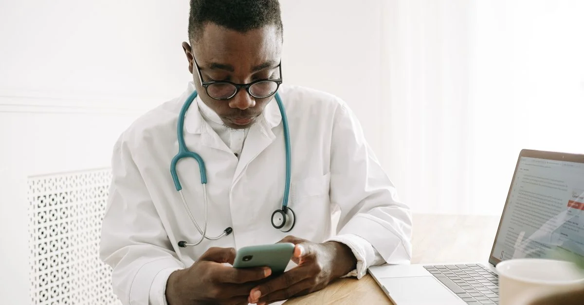 Médecin dans un cabinet médical vérifiant un smartphone avec un ordinateur portable sur la table, portant des lunettes et un stéthoscope.