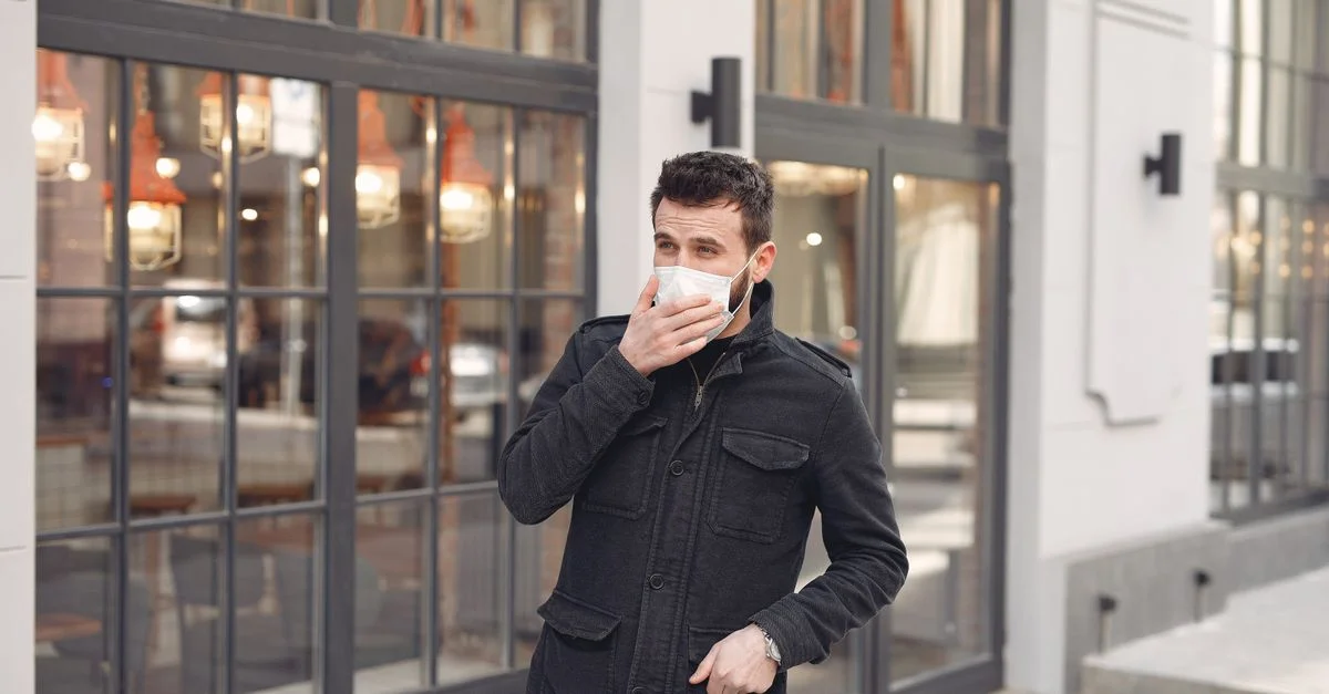 Homme en veste noire portant un masque facial dans une ville reflet de la normalité pandémique.