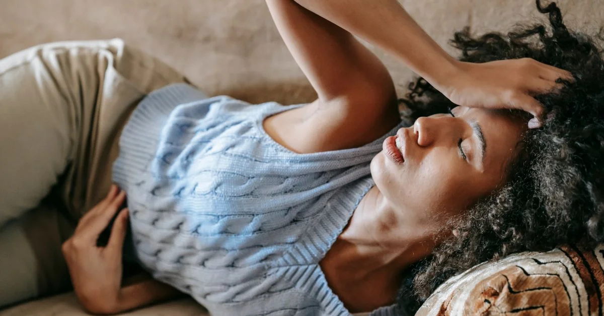 Une jeune femme aux cheveux bouclés est allongée à l'intérieur, ayant l'air troublée et touchant son front, peut-être malade ou souffrant d'un mal de tête.