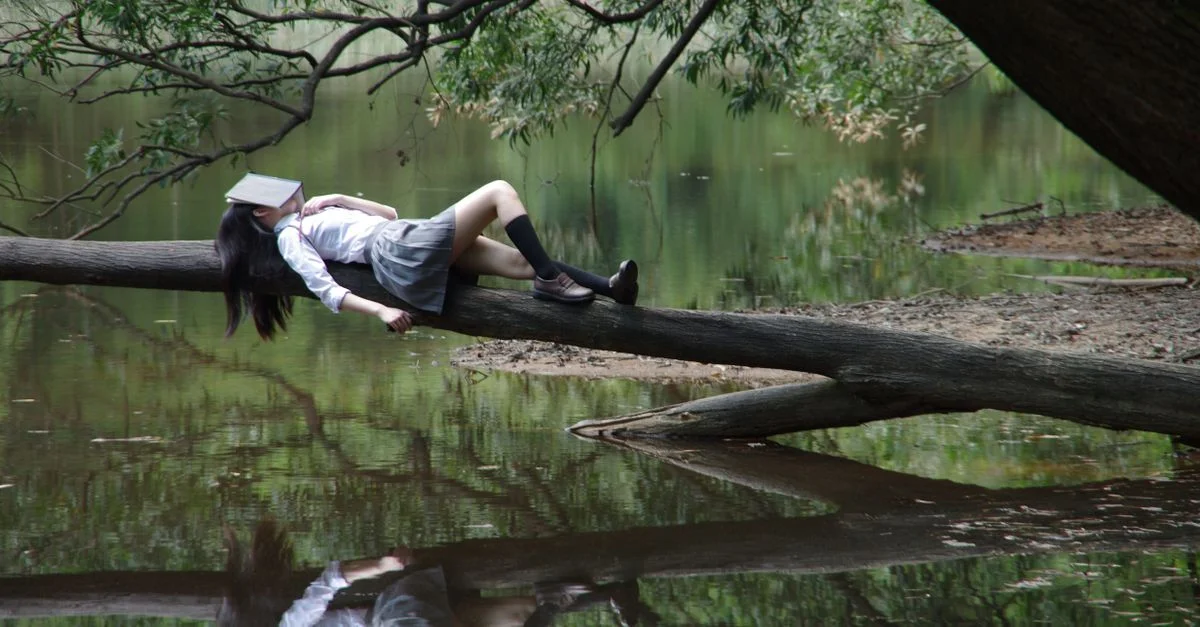 Une jeune femme avec un livre sur le visage se détend sur un tronc d'arbre au bord d'un lac tranquille.