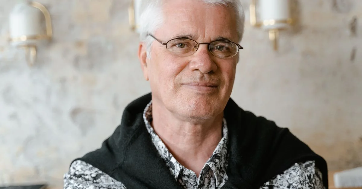 Portrait d'un homme senior souriant portant des lunettes et une chemise florale à l'intérieur.