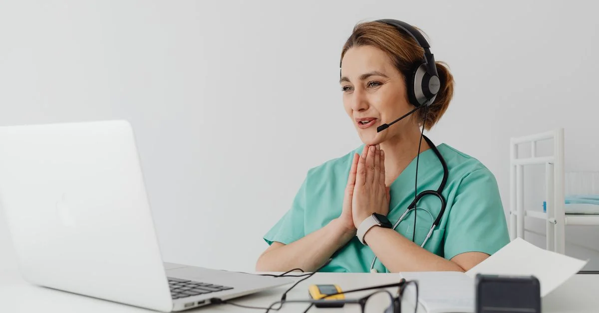 Infirmière utilisant la télémédecine pour une consultation virtuelle sur un ordinateur portable, portant des écouteurs et un stéthoscope.