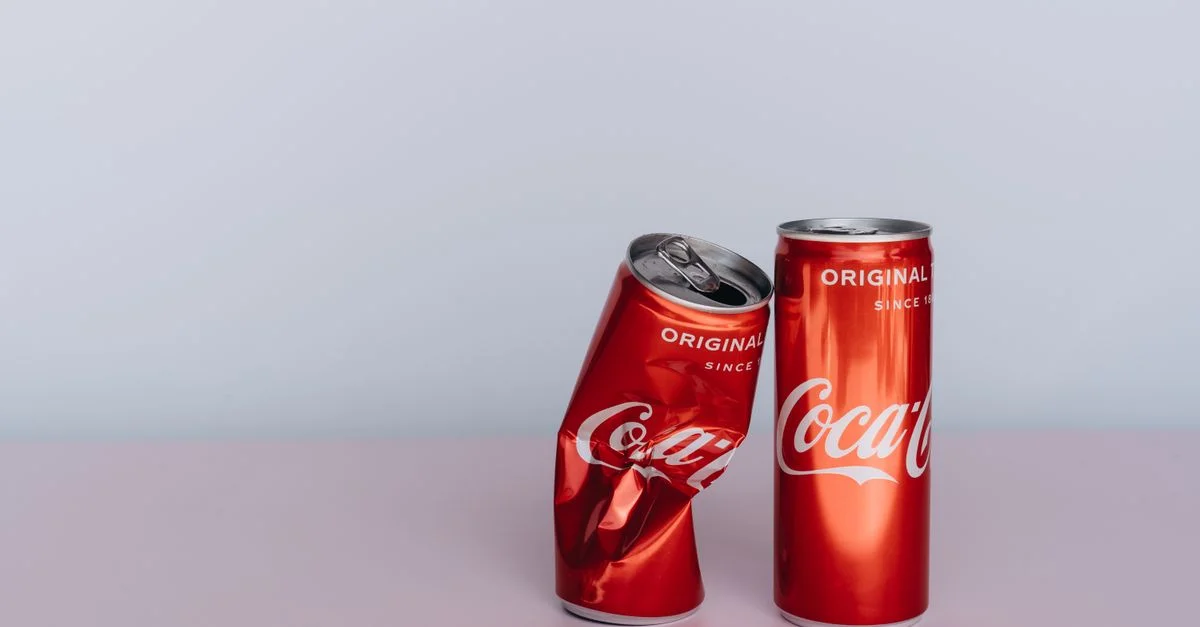 Deux canettes de Coca Cola, dont une froissée, sur une table minimaliste avec beaucoup d'espace pour le texte.