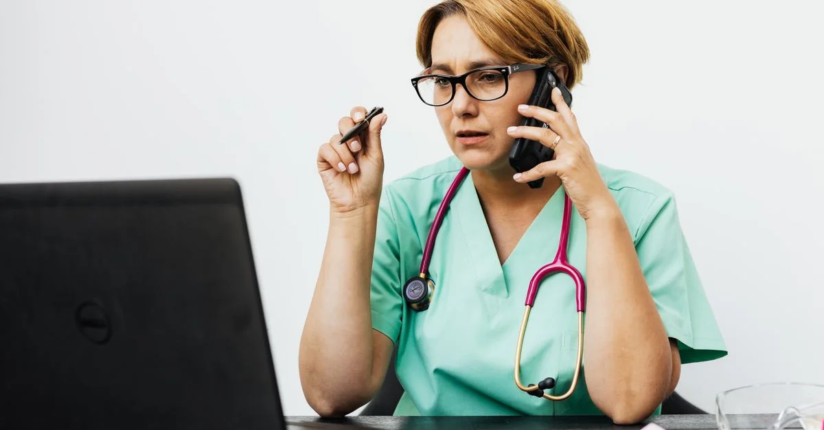 Une femme en tenue médicale travaillant avec un ordinateur portable et un téléphone, illustrant la communication moderne dans les soins de santé.