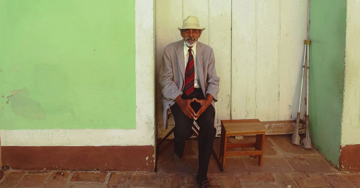 Homme senior avec un chapeau assis contre un mur vert, une canne à proximité en plein air.