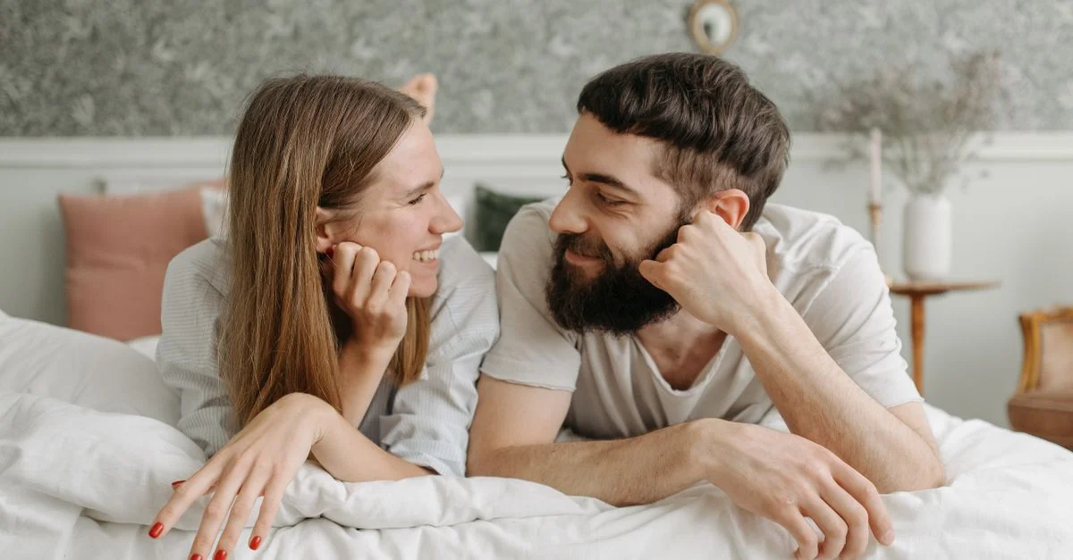 Couple heureux allongé face à face sur un lit, échangeant des sourires dans une chambre intime et chaleureuse.