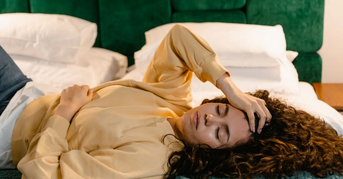 Femme allongée sur le lit avec la main sur le front, semblant détendue ou endormie dans un cadre intérieur confortable.